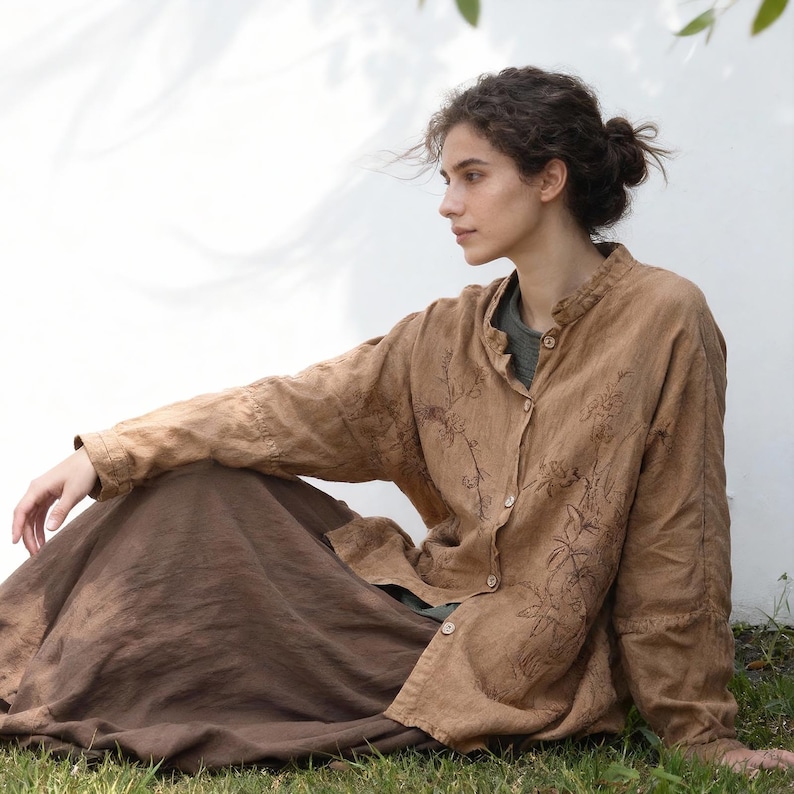 May include: A woman wearing a tan linen shirt with floral embroidery and a long brown skirt, sitting on the grass. The shirt is buttoned and has a mandarin collar. The outfit is casual and earthy.