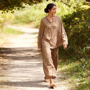 May include: A woman wearing a light brown linen outfit, including a button-down shirt with embroidered details and loose-fitting pants. The outfit is paired with woven sandals. The woman is walking on a dirt path surrounded by greenery.