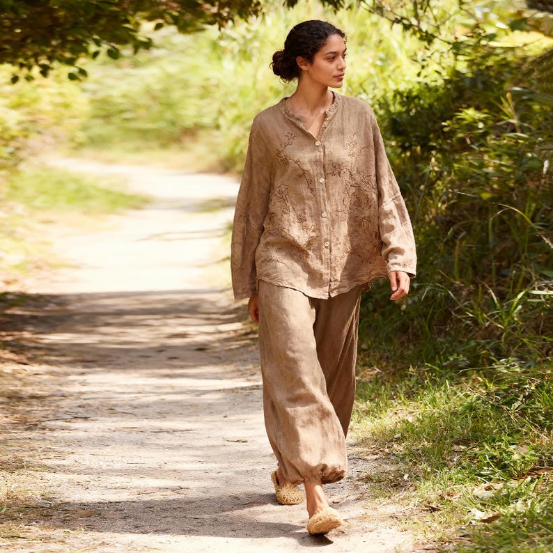 May include: A woman wearing a light brown linen outfit, including a button-down shirt with embroidered details and loose-fitting pants. The outfit is paired with woven sandals. The woman is walking on a dirt path surrounded by greenery.