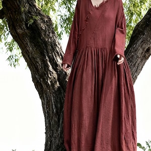 May include: A long, flowing, rust-coloured dress with long sleeves and a V-neck. The dress has a loose, comfortable fit and a gathered skirt. The fabric appears to be a soft, textured material. The dress is suitable for casual wear.