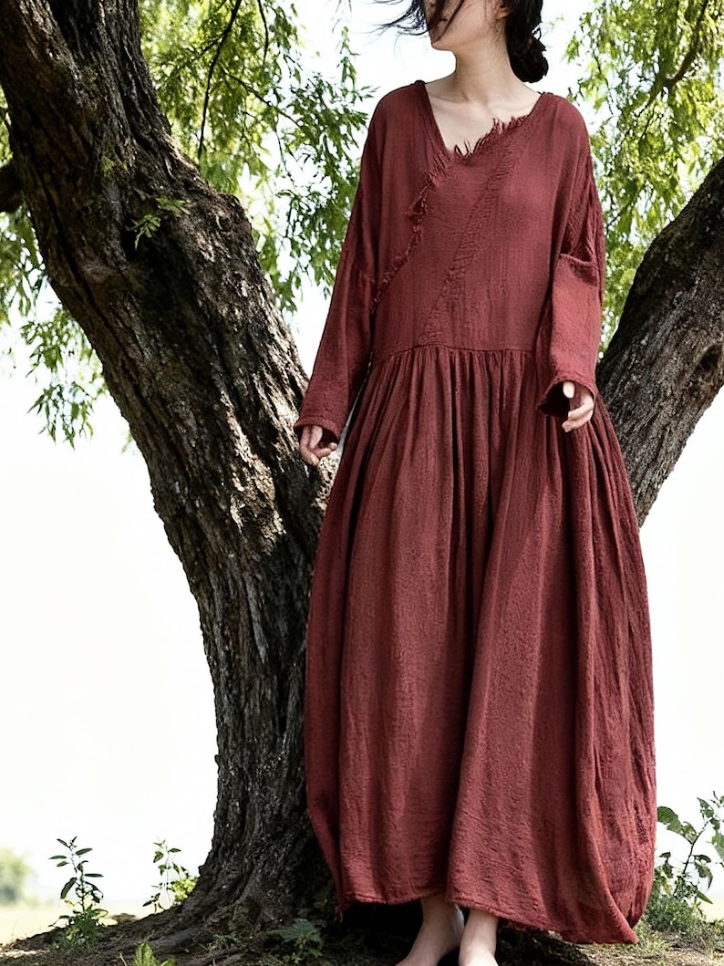 May include: A long, flowing, rust-coloured dress with long sleeves and a V-neck. The dress has a loose, comfortable fit and a gathered skirt. The fabric appears to be a soft, textured material. The dress is suitable for casual wear.