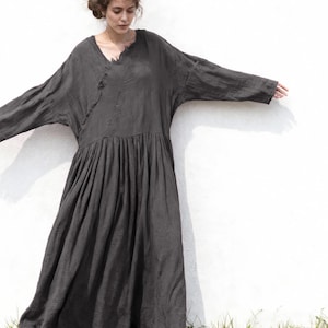 May include: A dark grey, long-sleeved dress with a V-neck and a gathered skirt. The dress has a loose, flowing silhouette and is made of a soft, textured fabric. The model is wearing sandals.