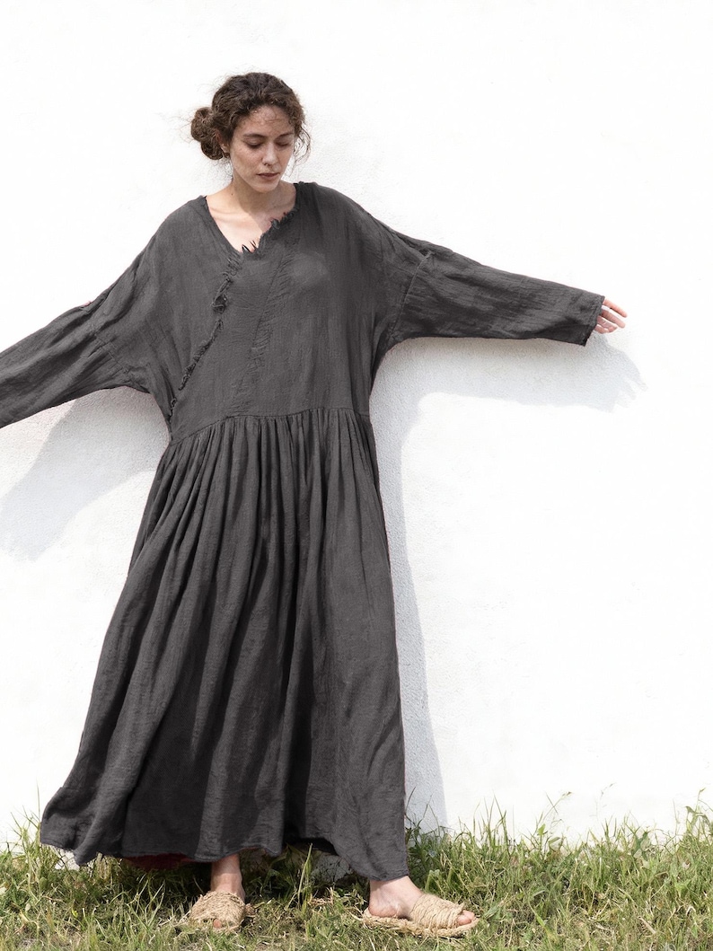 May include: A dark grey, long-sleeved dress with a V-neck and a gathered skirt. The dress has a loose, flowing silhouette and is made of a soft, textured fabric. The model is wearing sandals.