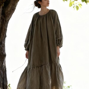 Puede incluir: Vestido largo verde oliva de corte holgado. Presenta mangas largas con pu&ntilde;os fruncidos, cuello redondo y falda escalonada con delicado ribete de encaje. La persona lleva el vestido al aire libre, cerca de un &aacute;rbol.