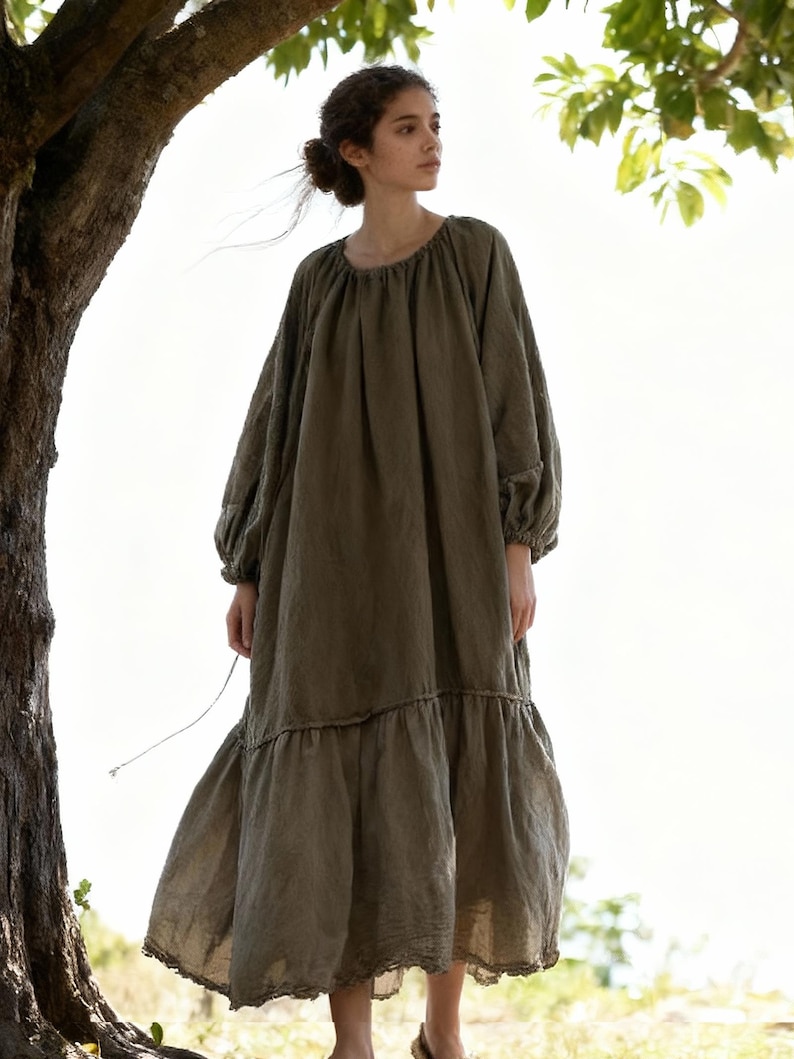 Puede incluir: Vestido largo verde oliva de corte holgado. Presenta mangas largas con pu&ntilde;os fruncidos, cuello redondo y falda escalonada con delicado ribete de encaje. La persona lleva el vestido al aire libre, cerca de un &aacute;rbol.