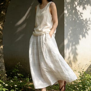 May include: A sleeveless, cream-colored linen top and a white plaid skirt. The skirt has a wide, flowing design with a subtle grid pattern. The outfit is worn outdoors in natural light, suggesting a casual, comfortable style.