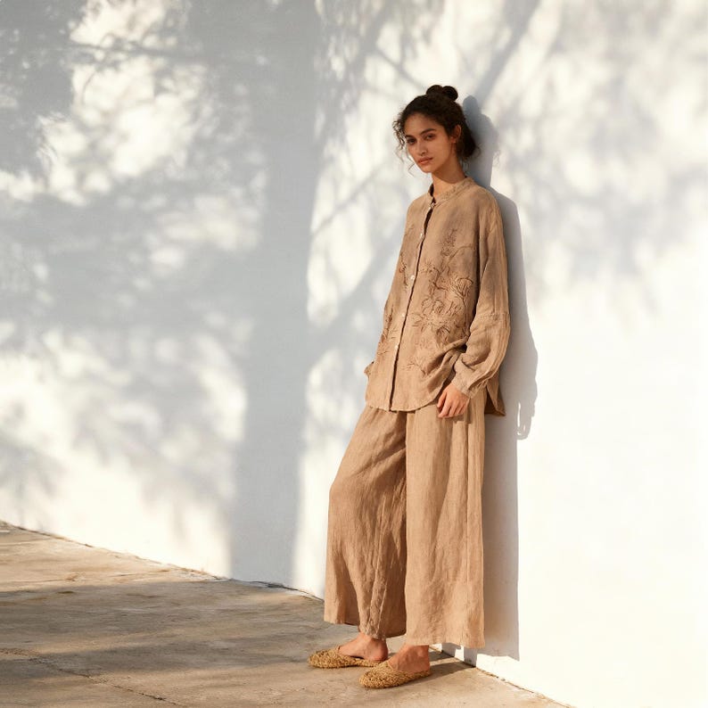 May include: A tan linen outfit, including a long-sleeved button-down shirt with floral embroidery and wide-leg pants. The outfit is paired with woven flats. The ensemble is a neutral color and appears to be lightweight.