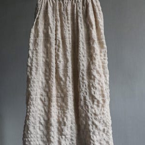 May include: A long, cream-colored skirt with a textured, puckered fabric. The skirt hangs from a hanger, showcasing its full length. Subtle, dark lines create a grid pattern across the fabric, adding visual interest to the garment.