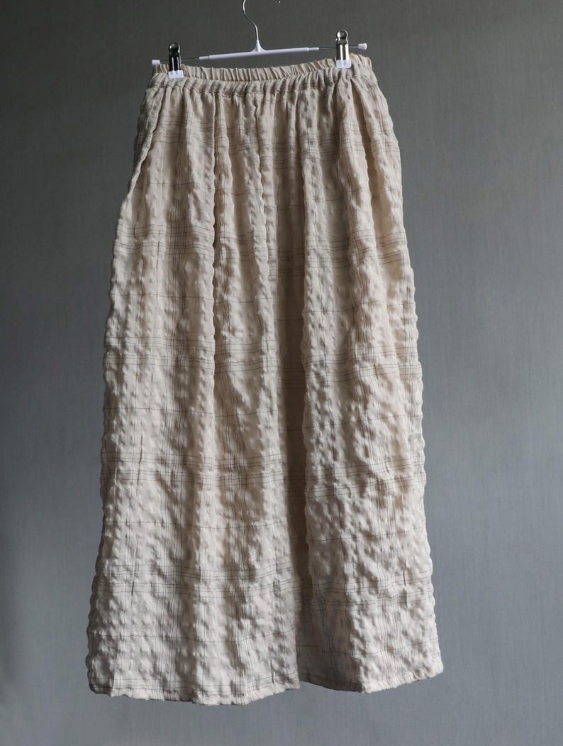 May include: A long, cream-colored skirt with a textured, puckered fabric. The skirt hangs from a hanger, showcasing its full length. Subtle, dark lines create a grid pattern across the fabric, adding visual interest to the garment.