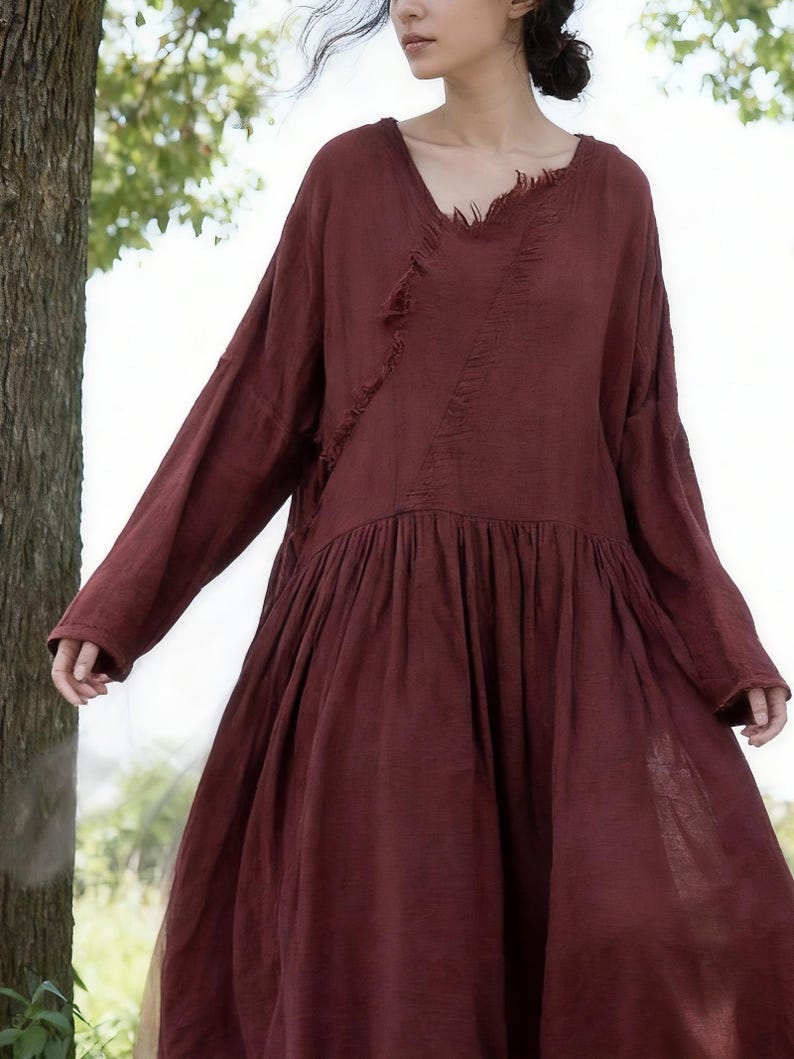 May include: A long-sleeved, burgundy dress with a V-neck and a gathered skirt. The dress has a raw-edge detail across the bodice and a flowing, relaxed fit. The dress is made of a soft, lightweight fabric.
