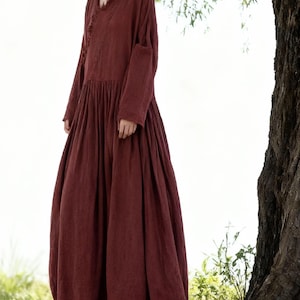 May include: A long, flowing burgundy dress with a V-neck and long sleeves. The dress has a gathered waist and a full, voluminous skirt. The fabric looks to be a soft, lightweight material. The dress is a solid colour with a simple, elegant design. The dress is suitable for warmer weather.