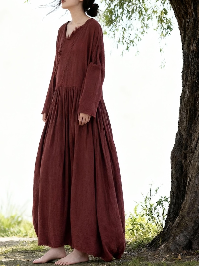 May include: A long, flowing burgundy dress with a V-neck and long sleeves. The dress has a gathered waist and a full, voluminous skirt. The fabric looks to be a soft, lightweight material. The dress is a solid colour with a simple, elegant design. The dress is suitable for warmer weather.