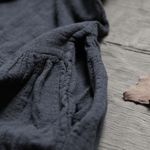 May include: Close-up of a dark grey garment with a textured, wrinkled fabric. The garment features a gathered pocket detail and a soft, flowing drape. A dried leaf rests on the grey surface next to the garment.