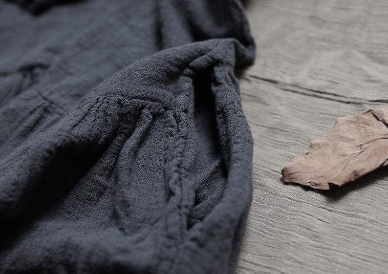 May include: Close-up of a dark grey garment with a textured, wrinkled fabric. The garment features a gathered pocket detail and a soft, flowing drape. A dried leaf rests on the grey surface next to the garment.