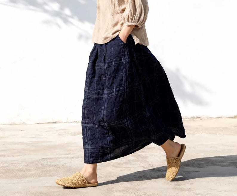 May include: A person wearing a beige linen top and a navy blue plaid skirt. The skirt is a flowing, wide-legged design. Woven, tan-colored slip-on shoes complete the outfit. The person is walking on a concrete surface.