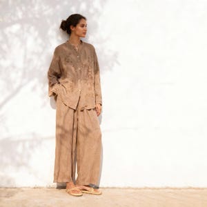May include: A woman wearing a light brown linen outfit, consisting of a long-sleeved button-down shirt with floral embroidery and wide-leg pants. The outfit is paired with woven sandals. The background is a white wall with shadows.