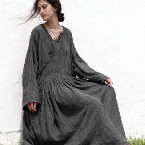 May include: A dark grey, long-sleeved dress with a V-neck and a gathered waist. The dress has a flowing, floor-length skirt and a relaxed fit. The model is sitting on the grass next to a white wall.