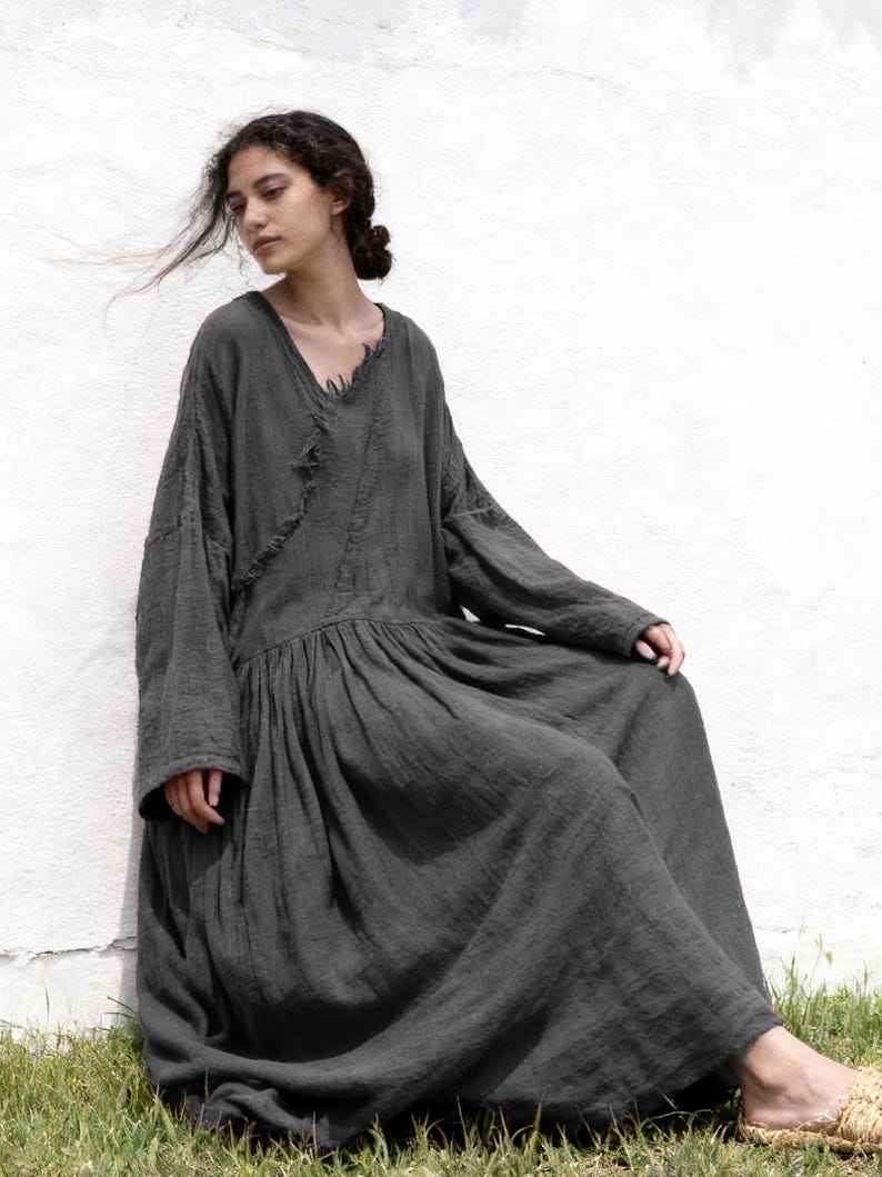 May include: A dark grey, long-sleeved dress with a V-neck and a gathered waist. The dress has a flowing, floor-length skirt and a relaxed fit. The model is sitting on the grass next to a white wall.