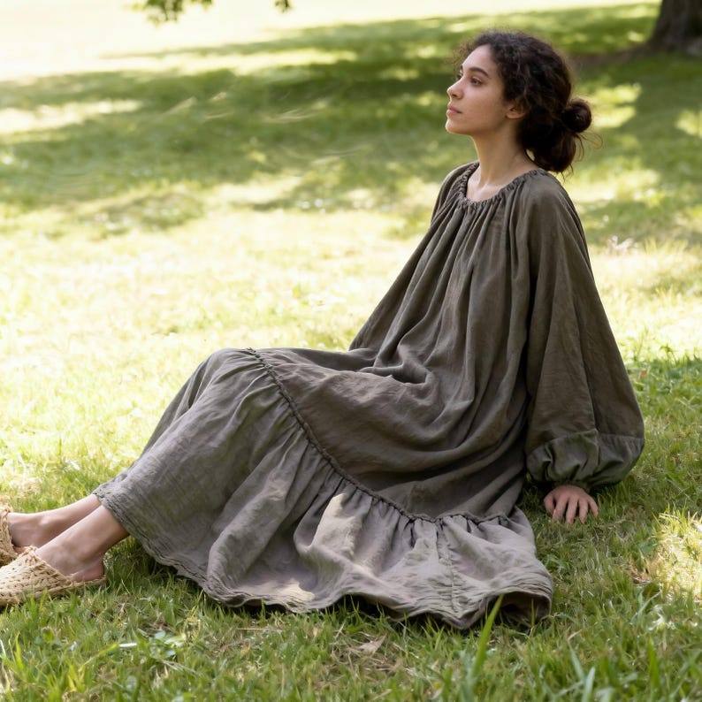 Puede incluir: Un vestido largo verde oliva con dobladillo con volantes y mangas largas y voluminosas. El vestido tiene un ajuste holgado y est&aacute; hecho de una tela suave y fluida. La persona est&aacute; sentada en la hierba en un parque.