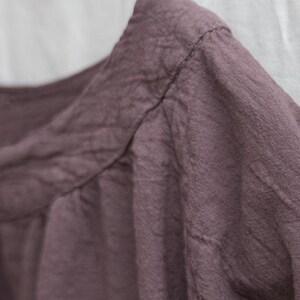 May include: Close-up of a dusty rose-coloured garment, possibly a shirt or blouse, with visible stitching along the neckline and shoulder. The fabric appears textured and slightly wrinkled, suggesting linen or similar material. The background is plain white.