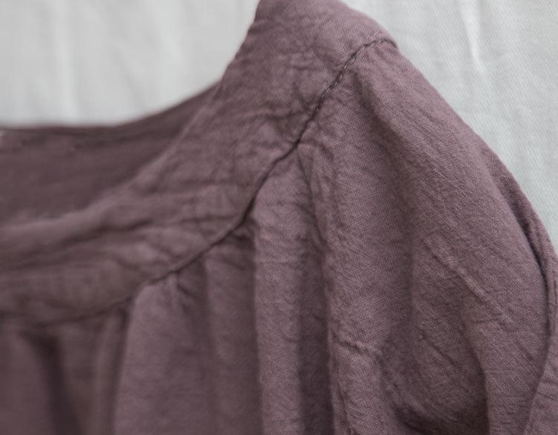 May include: Close-up of a dusty rose-coloured garment, possibly a shirt or blouse, with visible stitching along the neckline and shoulder. The fabric appears textured and slightly wrinkled, suggesting linen or similar material. The background is plain white.
