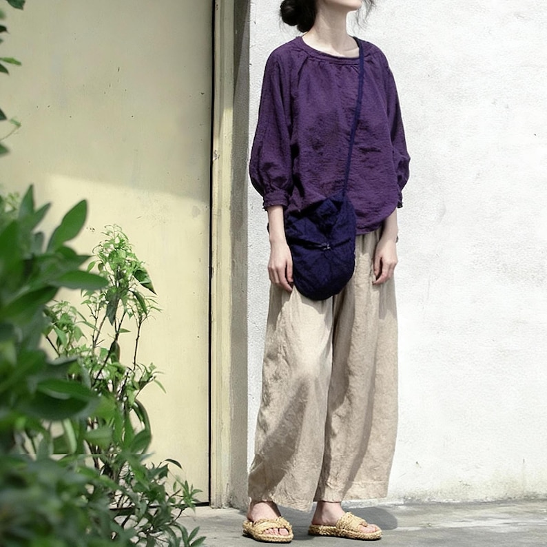 May include: An outfit comprising a purple long-sleeved blouse, tan wide-leg trousers, and a navy blue crossbody bag. The ensemble is finished with woven sandals. The blouse has a round neck and the bag features a quilted design.