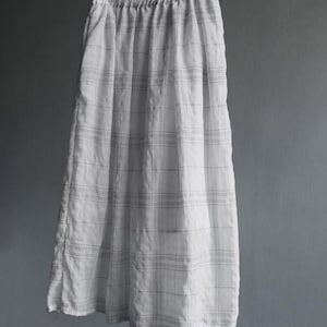 May include: A long, white skirt with a plaid pattern of thin, dark gray lines. The skirt is hanging from a metal hanger against a plain gray background. The fabric appears to be lightweight and slightly wrinkled.