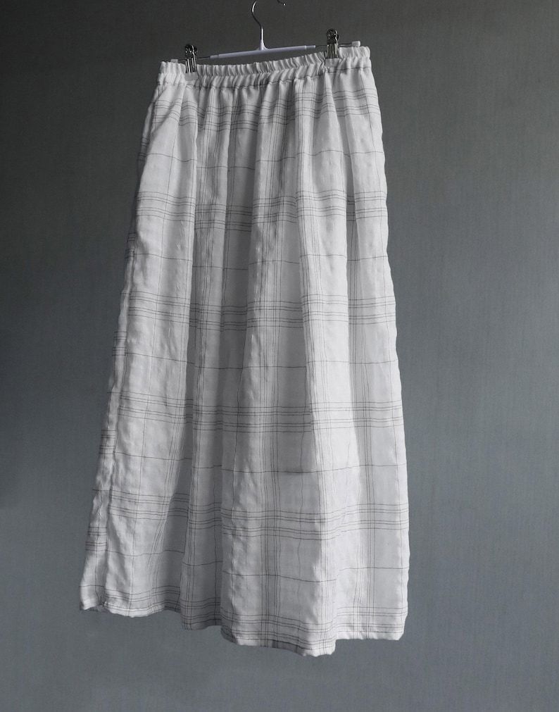 May include: A long, white skirt with a plaid pattern of thin, dark gray lines. The skirt is hanging from a metal hanger against a plain gray background. The fabric appears to be lightweight and slightly wrinkled.