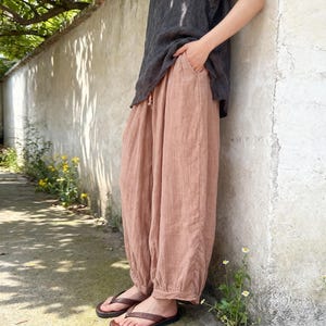 May include: A person wearing a dark gray top and light brown linen pants with a drawstring waist. The pants are loose-fitting with a gathered ankle. Brown leather flip-flops are worn on the feet.