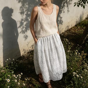 May include: A woman wearing a beige linen tank top and a white plaid skirt with pockets. The outfit is paired with brown sandals. The background features a white wall, a tree, and wildflowers.