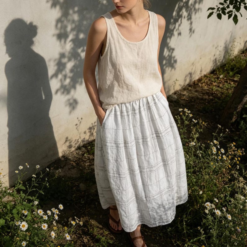 May include: A woman wearing a beige linen tank top and a white plaid skirt with pockets. The outfit is paired with brown sandals. The background features a white wall, a tree, and wildflowers.