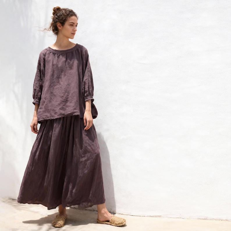 May include: A woman in a two-piece outfit. The outfit comprises a long-sleeved, scoop-neck top and a long, flowing skirt, both in a muted purple hue. She is also wearing woven sandals. The background is a white wall.