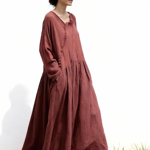 May include: A full-length, rust-coloured dress with long sleeves and a gathered skirt. The dress has a V-neckline with a fringed detail and pockets. The model is standing in front of a white wall and green grass.