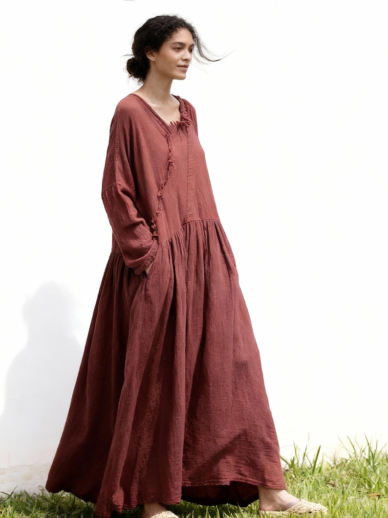 May include: A full-length, rust-coloured dress with long sleeves and a gathered skirt. The dress has a V-neckline with a fringed detail and pockets. The model is standing in front of a white wall and green grass.