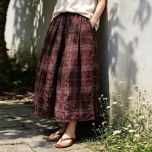 May include: A long, burgundy plaid skirt with a pleated design. The skirt is paired with brown sandals. The model is wearing a cream-colored top. The skirt has a relaxed fit and appears to be made of a lightweight fabric.