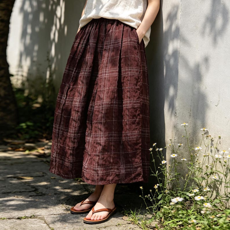 May include: A long, burgundy plaid skirt with a pleated design. The skirt is paired with brown sandals. The model is wearing a cream-colored top. The skirt has a relaxed fit and appears to be made of a lightweight fabric.