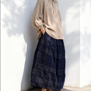 May include: A beige linen blouse with a tie-neck detail and long, voluminous sleeves. Paired with wide-leg, navy blue plaid pants. Woven, tan-colored slip-on shoes complete the outfit. The clothing is loose-fitting and casual.