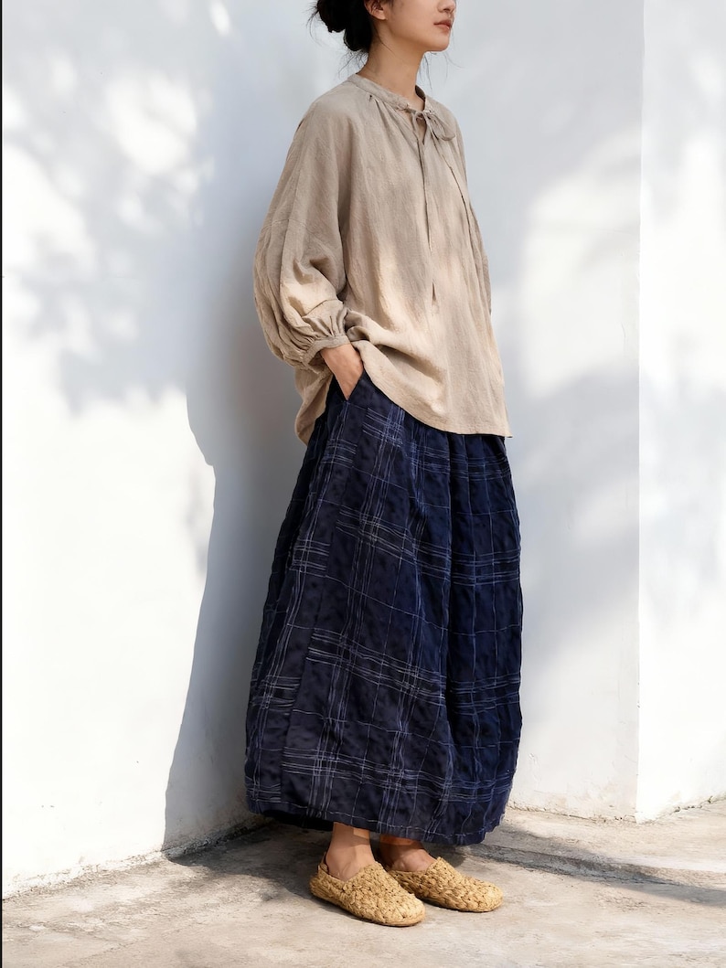 May include: A beige linen blouse with a tie-neck detail and long, voluminous sleeves. Paired with wide-leg, navy blue plaid pants. Woven, tan-colored slip-on shoes complete the outfit. The clothing is loose-fitting and casual.