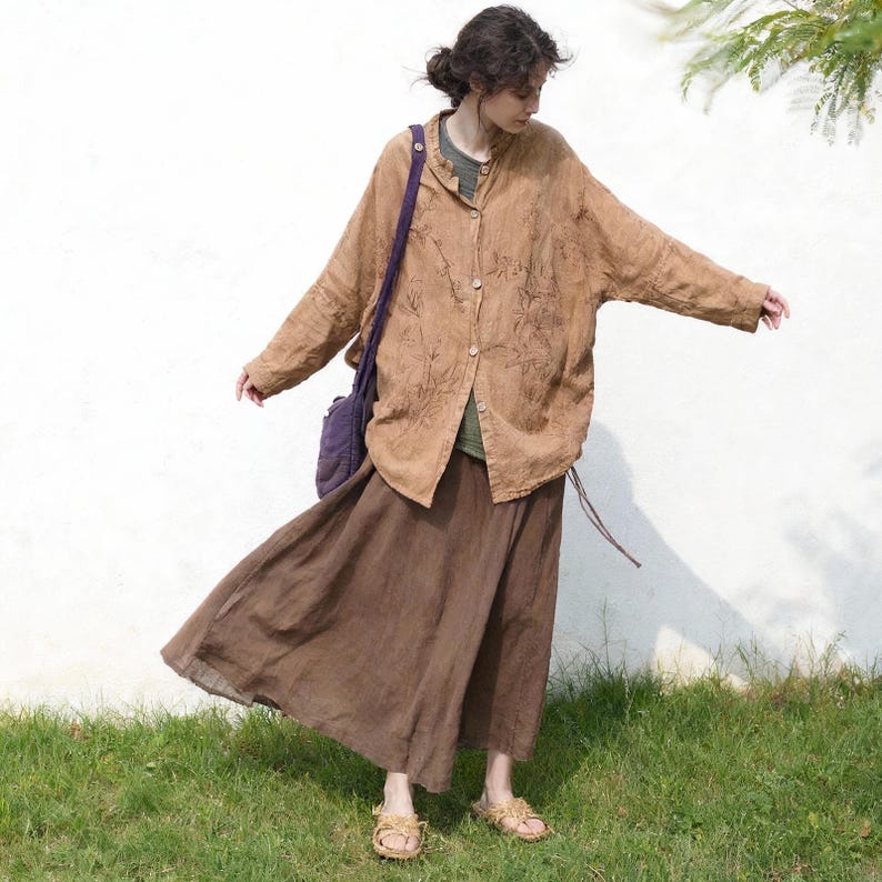 May include: A woman wearing a brown linen button-down shirt with embroidered details, a brown skirt, and a purple shoulder bag. The outfit is paired with sandals. The background is a white wall and green grass.