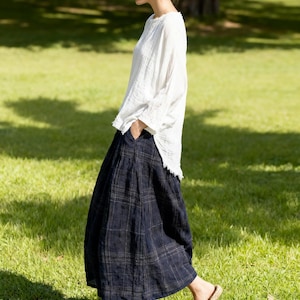 May include: A woman wearing a white long-sleeve top and a navy blue plaid skirt. She is walking on a grassy lawn, wearing woven flats. The outfit is casual and suitable for warm weather.