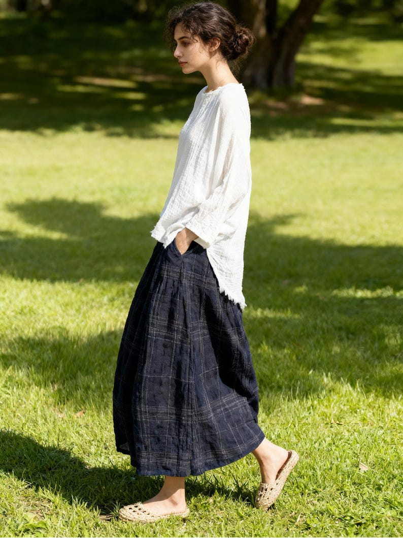 May include: A woman wearing a white long-sleeve top and a navy blue plaid skirt. She is walking on a grassy lawn, wearing woven flats. The outfit is casual and suitable for warm weather.