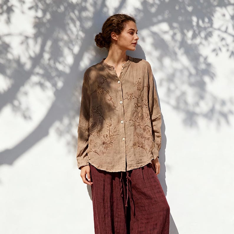 May include: A long-sleeved, button-down shirt in a light brown hue, adorned with embroidered floral patterns. The shirt is paired with burgundy-colored, wide-leg pants. The outfit is captured in natural light, creating a soft, warm aesthetic.