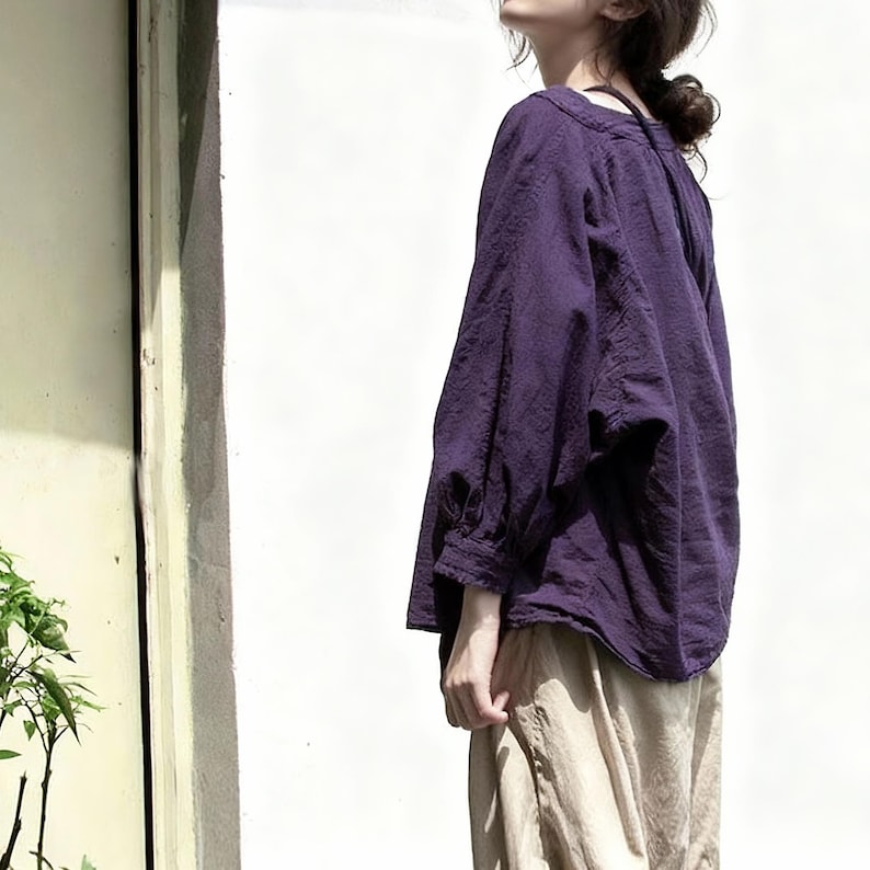 May include: A dark purple blouse with long sleeves and a round neckline. The blouse is worn with light beige trousers. The fabric has a textured appearance. The outfit is captured in natural light.