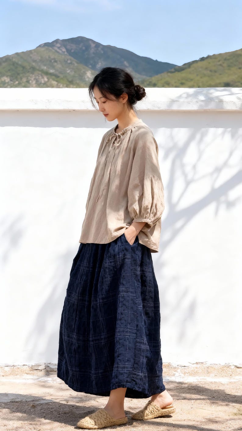 May include: A woman wearing a beige linen blouse with a tie-neck detail and three-quarter sleeves. She is wearing a long, navy blue plaid skirt. The outfit is completed with woven sandals. The background features a white wall and a mountain range.