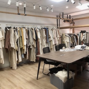 May include: A studio space featuring multiple metal clothing racks filled with various garments in neutral tones, including beige, white, and brown. A large wooden conference table with black chairs sits in the foreground, with a plastic bin containing fabric.