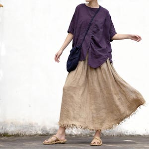 Puede incluir: Una blusa de manga corta color morado oscuro con una costura diagonal y detalle fruncido en el frente, combinada con una falda larga de color beige con textura, dobladillo deshilachado e irregular, bolso cruzado azul y sandalias trenzadas.