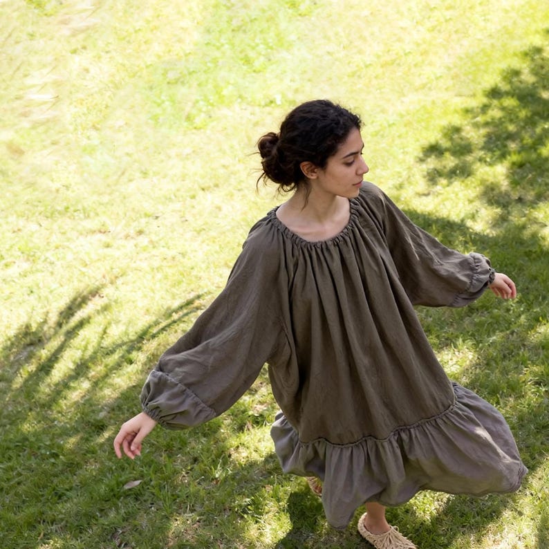 Puede incluir: Vestido verde oliva de manga larga, con cuello fruncido y dobladillo con volantes. El vestido tiene una silueta suelta y fluida y est&aacute; hecho de una tela suave y ligera. La persona est&aacute; de pie sobre c&eacute;sped.