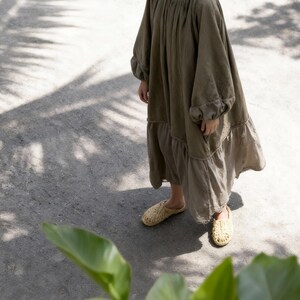 Puede incluir: Un vestido largo verde oliva con mangas largas y dobladillo con volantes. El vestido se combina con zapatos sin cordones beige tejidos. La imagen est&aacute; tomada al aire libre, con sombras de follaje en el suelo.