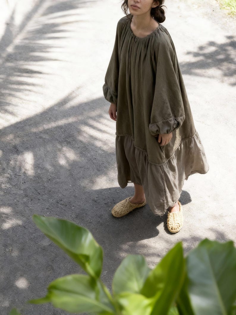 Puede incluir: Un vestido largo verde oliva con mangas largas y dobladillo con volantes. El vestido se combina con zapatos sin cordones beige tejidos. La imagen est&aacute; tomada al aire libre, con sombras de follaje en el suelo.