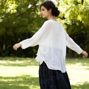 May include: A white, long-sleeved blouse with a loose fit and a scoop neckline. The blouse is paired with a dark blue plaid skirt. The image is taken outdoors with a green background.